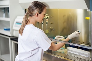Frau in einem Labor nimmt hinter einer Glasscheibe eine Probe mit einer Pipette aus Reagenzglas