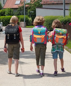 Trois enfants, dont un avec un cartable noir peu visible et deux avec des cartables en couleur bien visibles avec des éléments réfléchissants et fluorescents