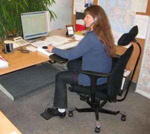 Frau arbeitet am inklusiv angepassten Bildschirmarbeitsplatz mit Headset, angepasstem Bürostuhl und Fußtaster.