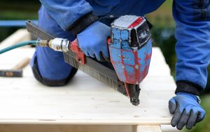 Person in Arbeitskleidung befestigt mit einem Druckluftnagler ein Brett auf einem Balken