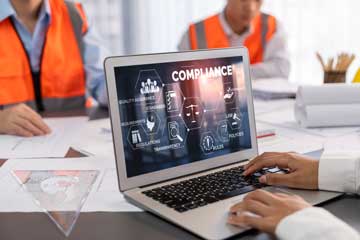 Ein aufgeklappter Laptop zeigt einen Bildschirm mit dem Wort Compliance und mehreren dazugehörigen Symbolen wie Regeln, Qualitäts­sicherung und Standards. Gegenüber am Tisch sitzen zwei Personen in orangefarbenen Warnwesten, die Bauzeichnungen vor sich liegen haben.. Klick öffnet die Seite Anwendungsbeginn der Bauprodukteverordnung