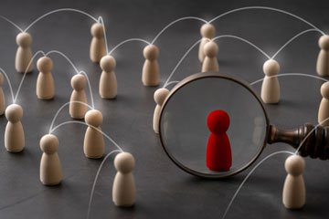 Verschiedene helle Holzpuppen stehen mit Abstand beisammen, darunter eine rote Holzpuppe, die durch eine Lupe betrachtet wird.. Klick öffnet die Seite DGUV Risikoobservatorium zur frühzeitigen Erkennung von Risiken in der Arbeitswelt