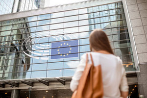 Junge Geschäftsfrau steht vor der Glasfront eines Gebäudes, in der sich die europäische Flagge spiegelt.
