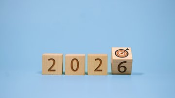 Holzwürfel bilden die Zahl 2026. Der letzte Würfel mit der 6 ist leicht gekippt, auf der angrenzenden Würfelfläche sieht man eine Zielscheibe. 