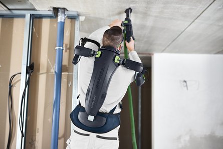 A man wearing an exoskeleton supporting his arms is drilling a hole in the ceiling
