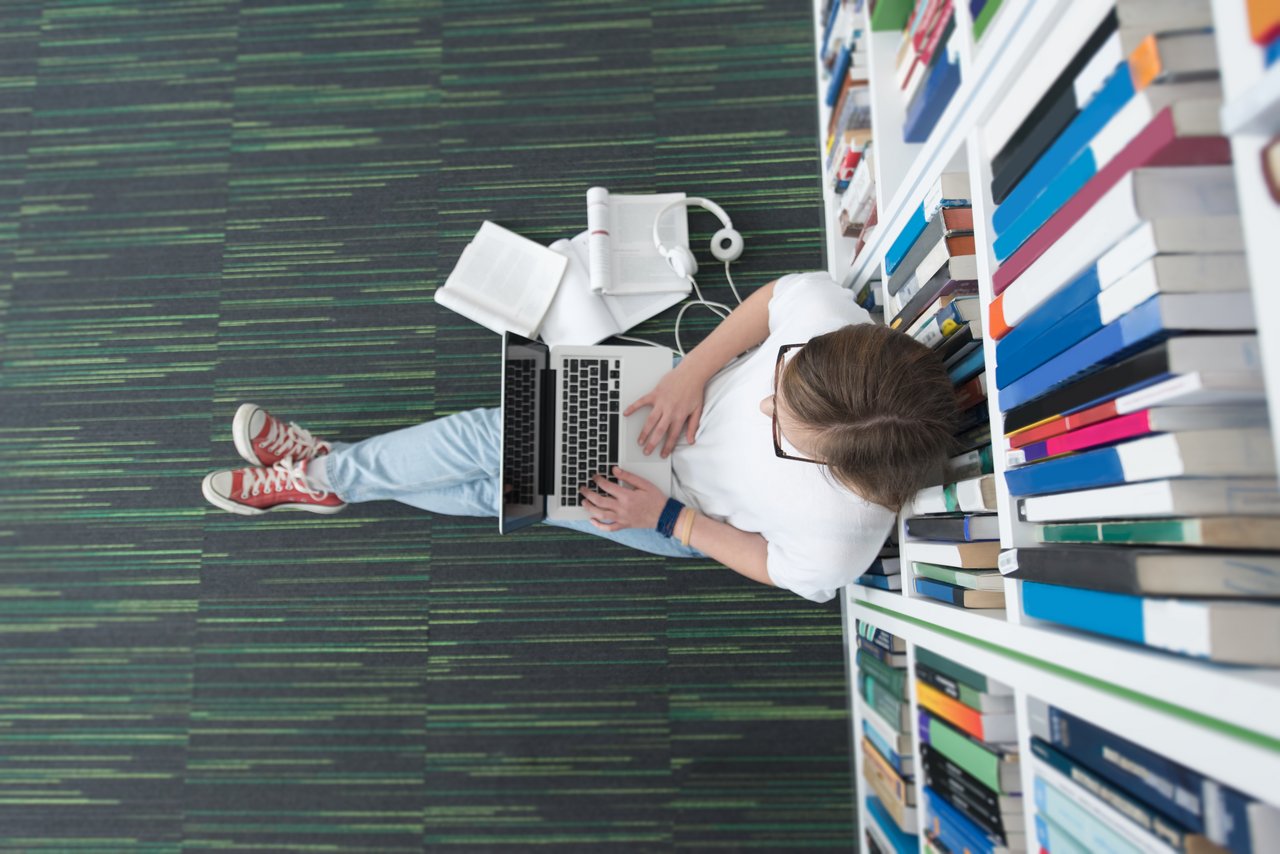 Studentin mit Laptop auf dem Schoß, vor einem Bücherregal auf dem Boden sitzend