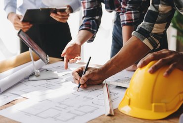 Drei Personen sind über einen Bauplan gebeugt und zeigen auf verschiedene Stellen. Neben dem Bauplan liegen Papierrollen und ein gelber Schutzhelm.. Klick öffnet die Seite KAN äußert sich zu Plänen für einen Rechtsakt für Baudienstleistungen