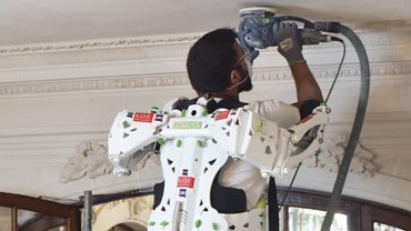 Ein Handwerker schleift die Decke eines Raumes. Er trägt ein Exoskelett, das ihn beim Halten der Maschine unterstützt.
