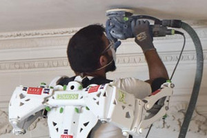 Ein Handwerker schleift die Decke eines Raumes. Er trägt ein Exoskelett, das ihn beim Halten der Maschine unterstützt.