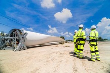 Ingenieure in gut sichtbarer Schutzkleidung begutachten auf einer Baustelle unter strahlend blauem Himmel ein großes Windrad-Rotorblatt