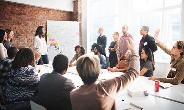 Gruppe von Menschen in einem Seminar, einige Personen mit erhobenen Händen, vorne ein Pinnwand mit Zetteln