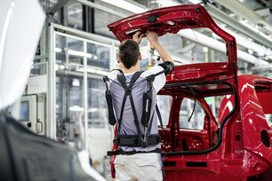 Mann mit Exoskelett arbeitet an geöffneten Heck eines Fahrzeuges in Fabrikhalle