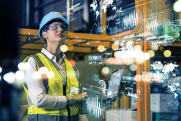 Eine Beschäftigte in Sicherheitskleidung sieht durch eine Sicherheitsbrille in eine durch augmented reality vernetzte Arbeitswelt 
