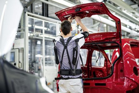 Dans une usine automobile, un homme portant un exosquelette pour soutenir ses bras est en train de monter quelque chose sur le hayon d'un coffre.