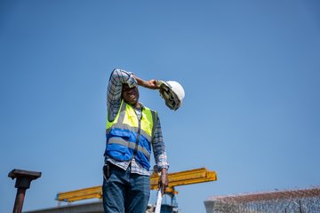 Bauarbeiter auf einer Baustelle wischt sich mit Helm in der Hand die Stirn ab; blauer Himmel und Sonnenschein