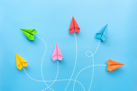 Sechs bunte Papierflieger auf blauem Hintergrund; gestrichelte Linien deuten verschiedene Flugrichtungen an