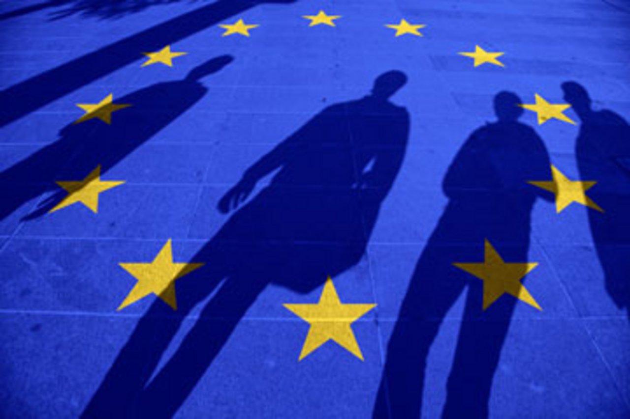 Mehrere Personen werfen Schatten auf Boden, auf den eine Europa-Flagge projiziert ist. 